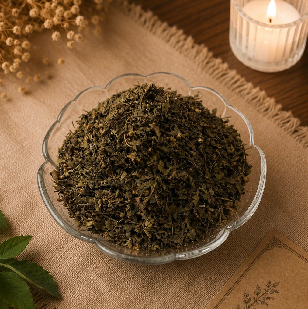 A glass bowl filled with loose dried green tea leaves, styled on a burlap runner with a lit candle, dried flowers, and a Tulsi leaf in the background.