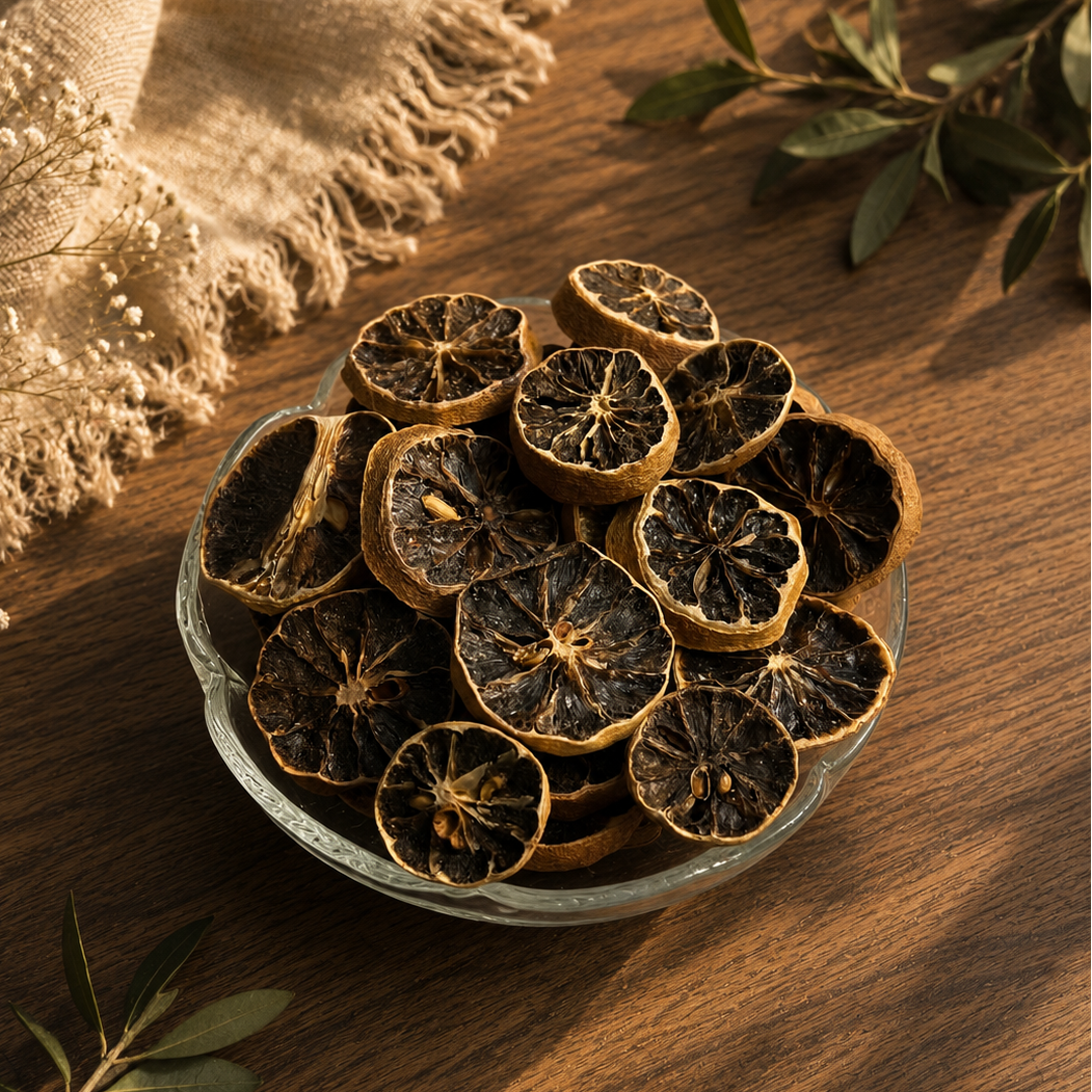 Dried citrus slice chips in a small clear glass bowl, arranged on a warm wooden surface with a beige fringed cloth and green leaves as styling props.