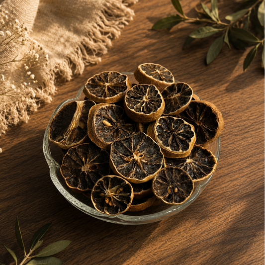 Dried citrus slice chips in a small clear glass bowl, arranged on a warm wooden surface with a beige fringed cloth and green leaves as styling props.