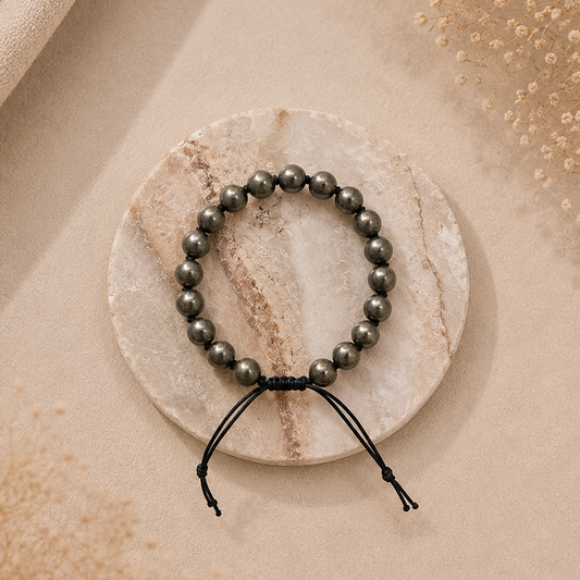 A beaded bracelet with dark gray metallic beads and a black adjustable cord, displayed on a round white marble coaster over a beige fabric background.
