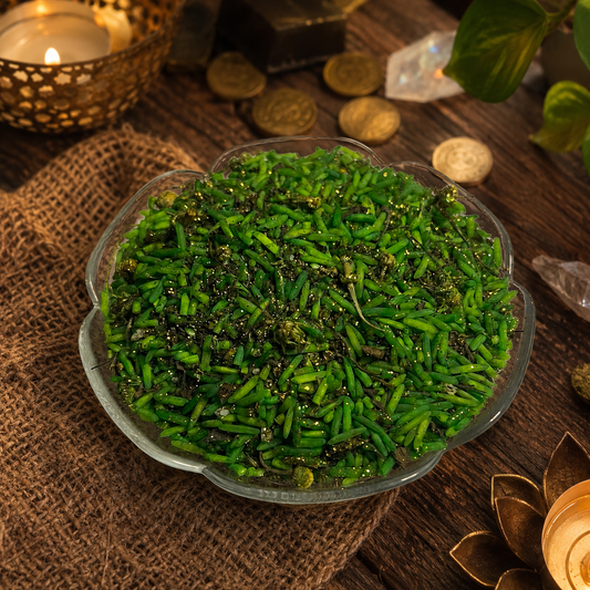 A clear glass bowl filled with chopped bright green herbs or scallions mixed with dark spices, set on a rustic wooden table with burlap, coins, crystals, and a lit candle in the background.