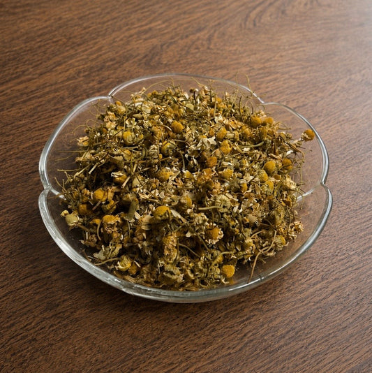 Dried chamomile tea mixture in a small clear glass bowl, placed on a dark brown wooden tabletop.