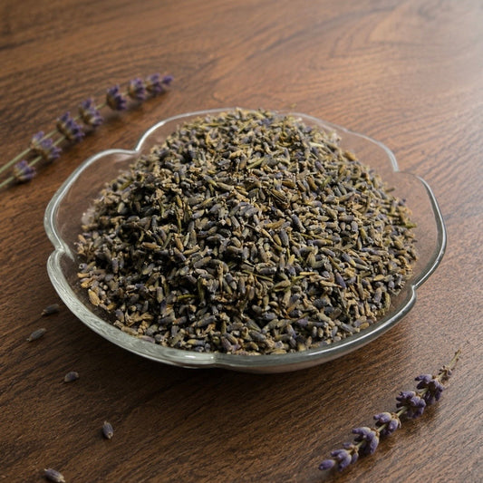 Dried lavender buds in a small clear glass bowl on a wooden surface, with a few sprigs placed around the bowl.