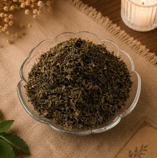 A glass bowl filled with loose dried green tea leaves, styled on a burlap runner with a lit candle, dried flowers, and a Tulsi leaf in the background.