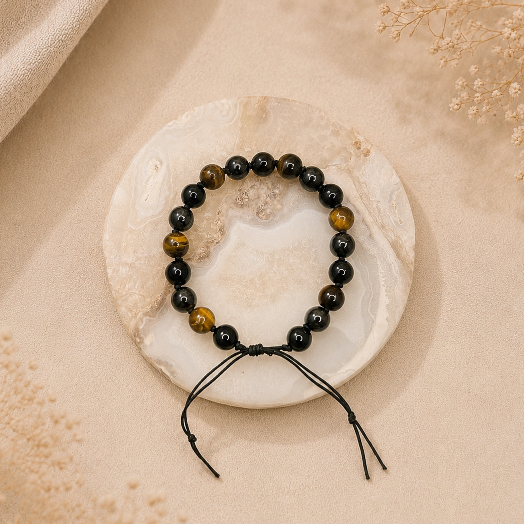 A beaded bracelet with black and brown tiger's eye-like beads and black cord ties, displayed on a round white stone coaster over a beige fabric background.