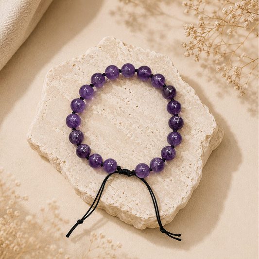 A beaded stretch bracelet made of polished purple gemstone beads with a black cord, displayed on a textured light beige stone slab with dried floral accents.